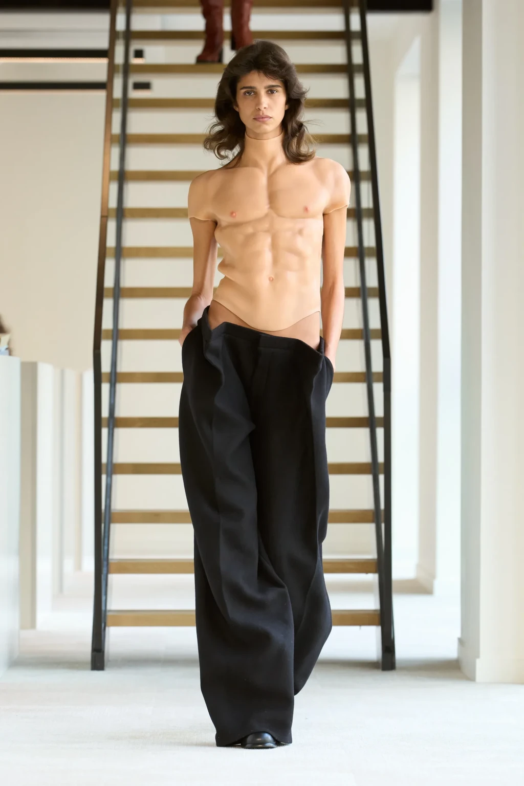 Fashion model walks the runway wearing black pants and a tan muscle suit, with a staircase in the background during a fashion show.