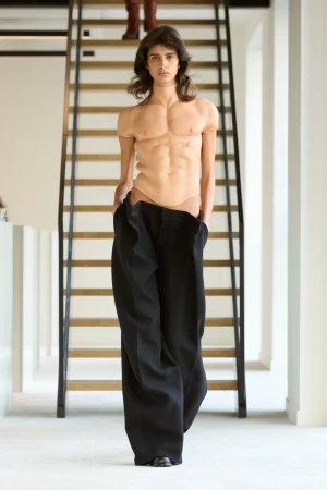 Fashion model walks the runway wearing black pants and a tan muscle suit, with a staircase in the background during a fashion show.