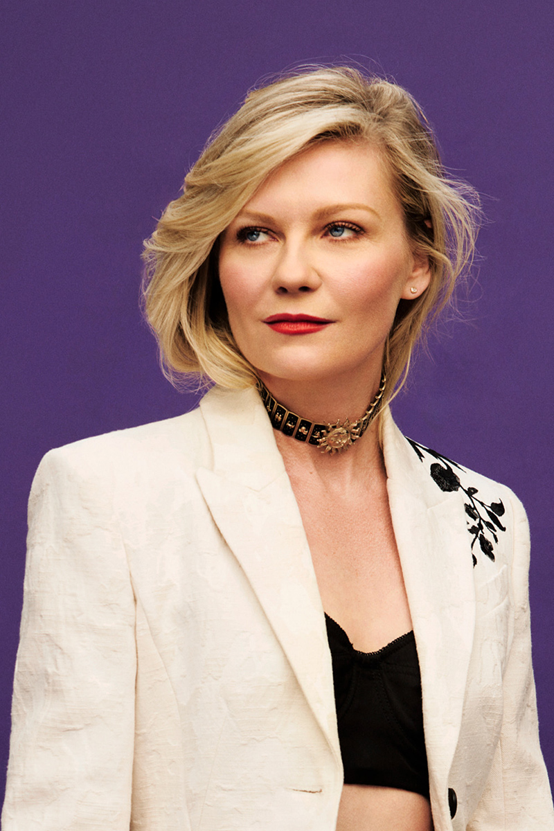 Kirsten Dunst in a studio portrait, wearing a white blazer and black choker against a purple background. Professional headshot.