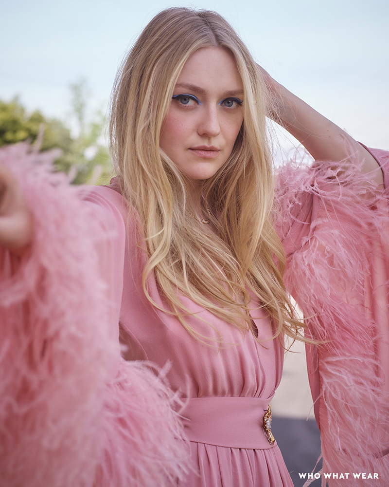 Blonde woman, Dakota Fanning, poses in pink feathered dress against a neutral outdoor background, showcasing a blend of fashion and nature.