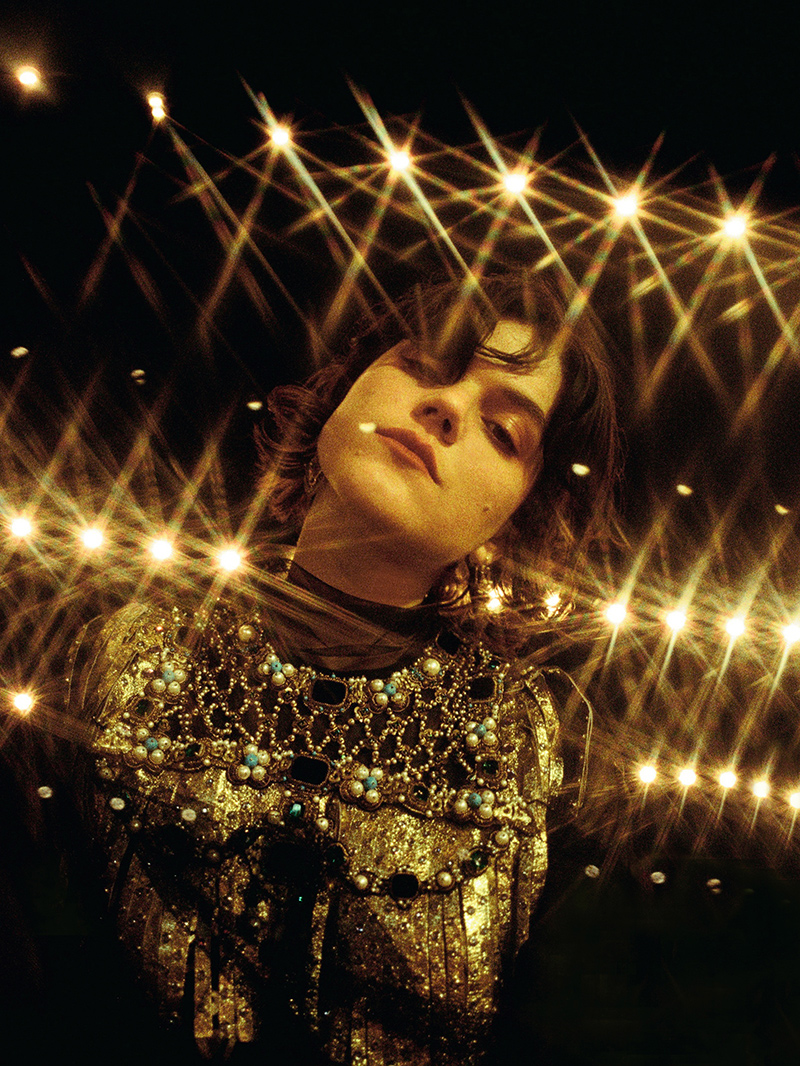 A young adult female gazes upward, illuminated by warm, golden bokeh lights, wearing an ornate gold top with jewel embellishments.