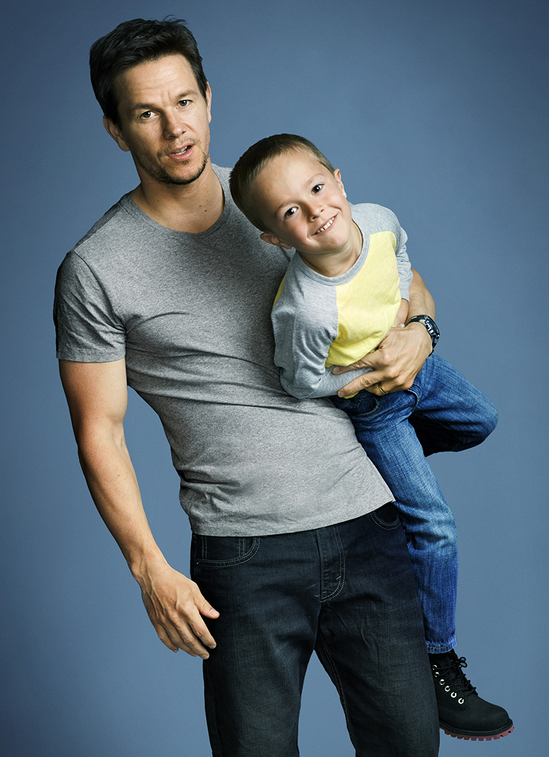 Mark Wahlberg holds a young child in a studio shot. The photo features a simple blue background and showcases the bond between the two.