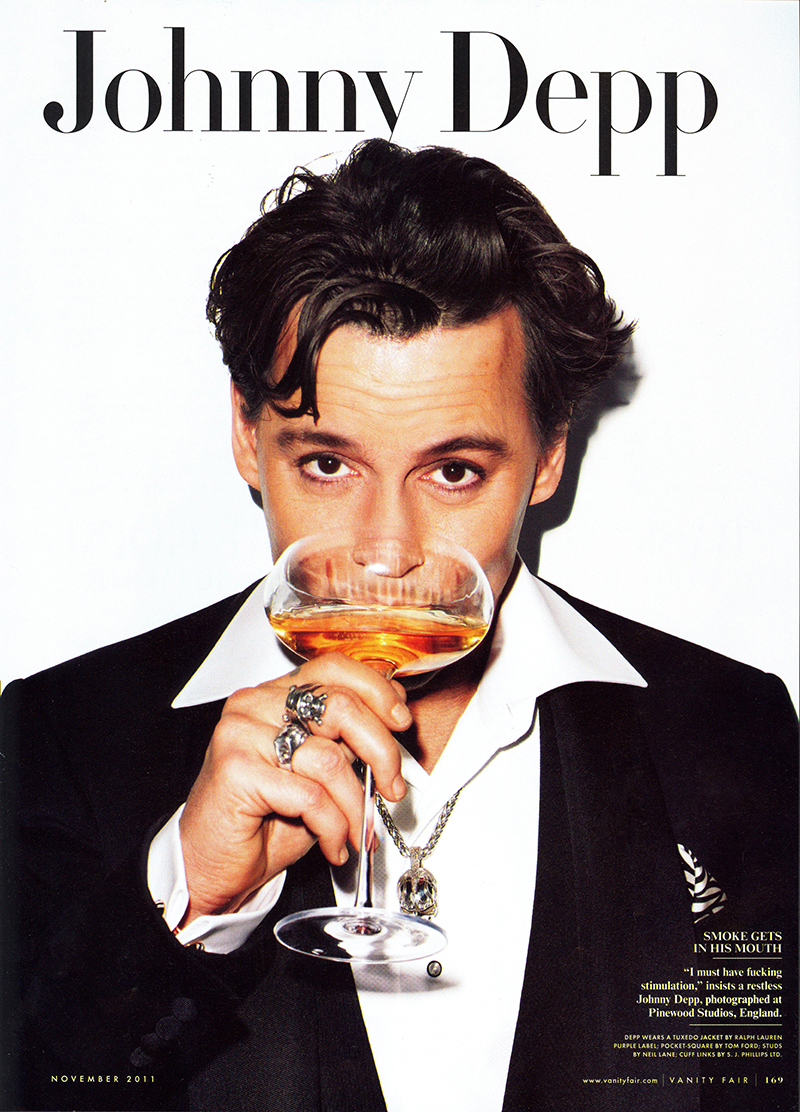 Johnny Depp portrait on a magazine cover; he holds a glass with a brown liquid. He is wearing a black suit, rings, and a necklace.