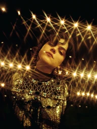 Woman in an ornate metallic gold embellished garment beneath glittering starburst string lights at night.