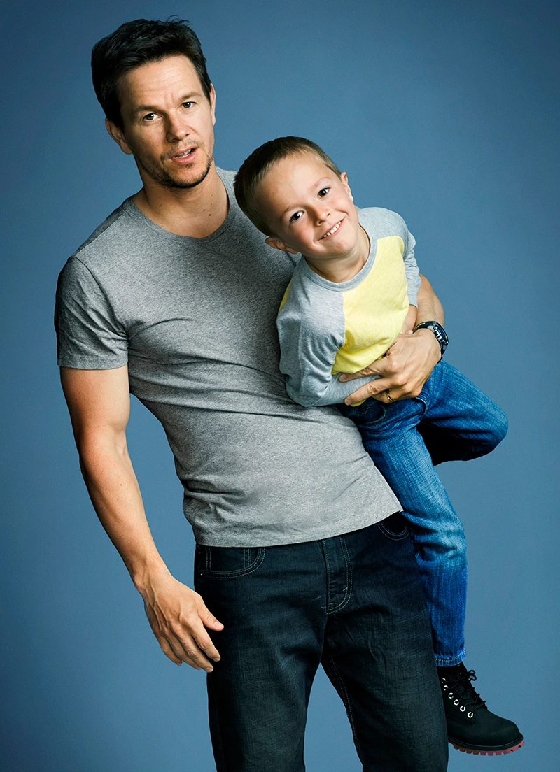Mark Wahlberg holding a young child in a studio portrait against a blue backdrop