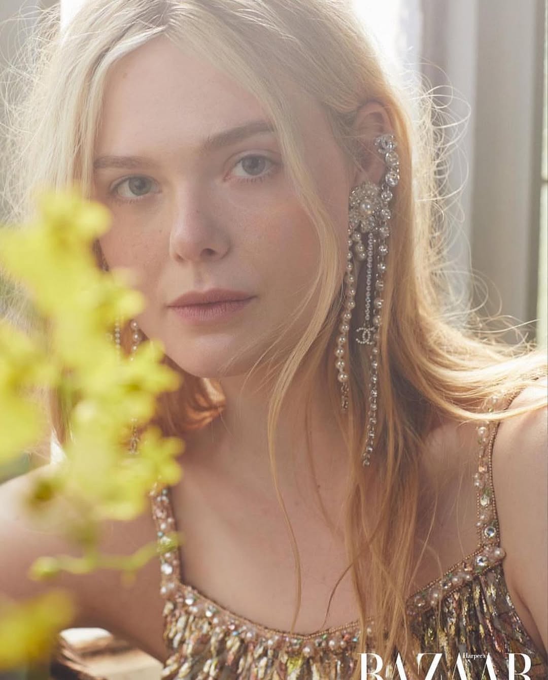 Close-up portrait of Elle Fanning with blond hair wearing a beaded and jeweled dress, posing with statement earrings, shot indoors in soft lighting.