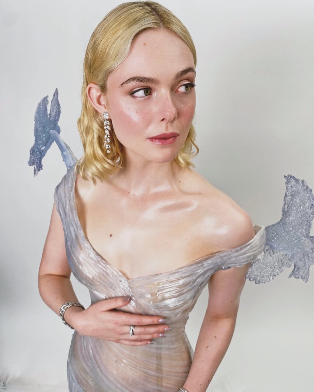 Elle Fanning poses in an elegant, light silver gown with decorative details, accessorized with jewelry against a plain white backdrop.