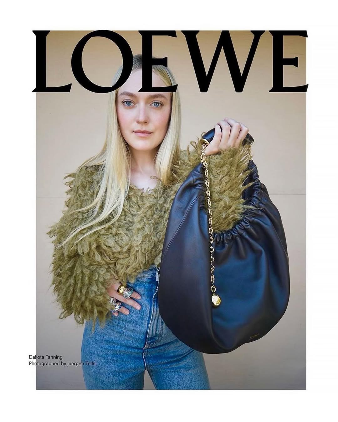 Dakota Fanning poses with a black leather Loewe purse featuring a gold chain, wearing a shaggy sweater and blue jeans against a plain background.