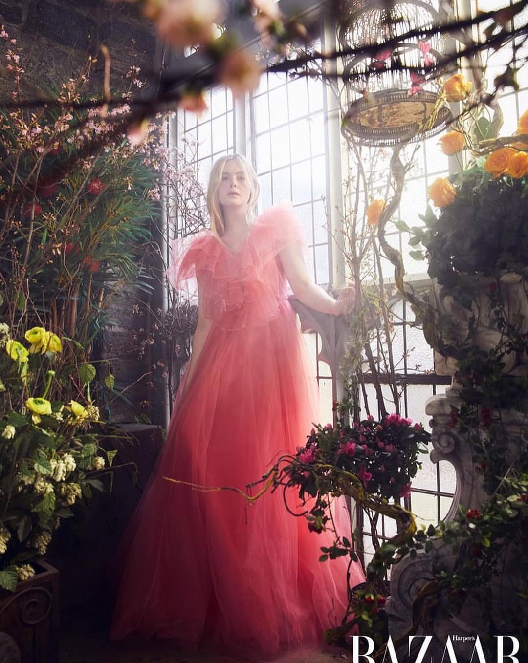 Woman in a pink gown poses among flowers, next to a stone staircase and birdcage in a light-filled, overgrown greenhouse.