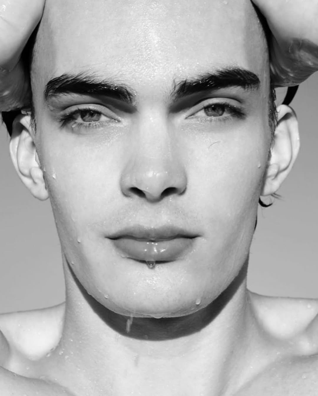 Close-up black and white portrait of a young adult man with water droplets on his face, head tilted back, and hands near his head.