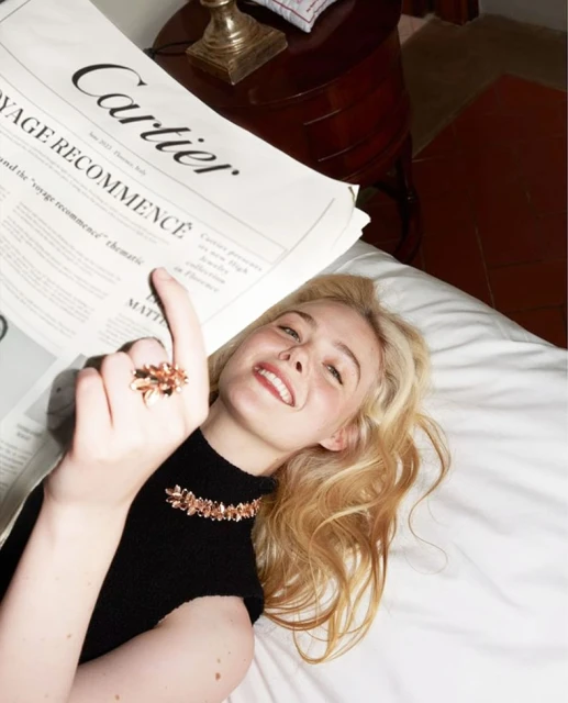 Blonde woman lying on white bed with Cartier newspaper, wearing necklace and ring with flower design, smiling with fair skin.