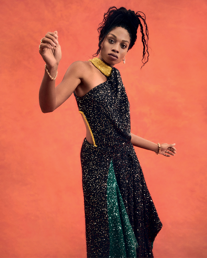 Adult female in a one-shoulder sequin gown with a gold collar and bracelets poses against an orange background in a studio shot.
