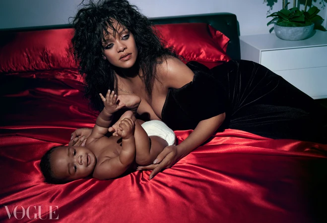 Editorial fashion shot of Rihanna in a black velvet dress lying on a red satin bed next to her infant child, Vogue magazine logo visible.