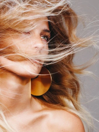 Close-up portrait of a woman with blonde hair blowing across her face and a large round gold hoop earring against a grey backdrop.