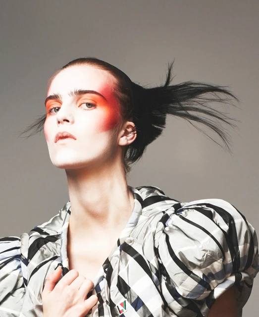 Fashion portrait of a female model with striking orange eye makeup and a black and white patterned jacket in a studio setting.