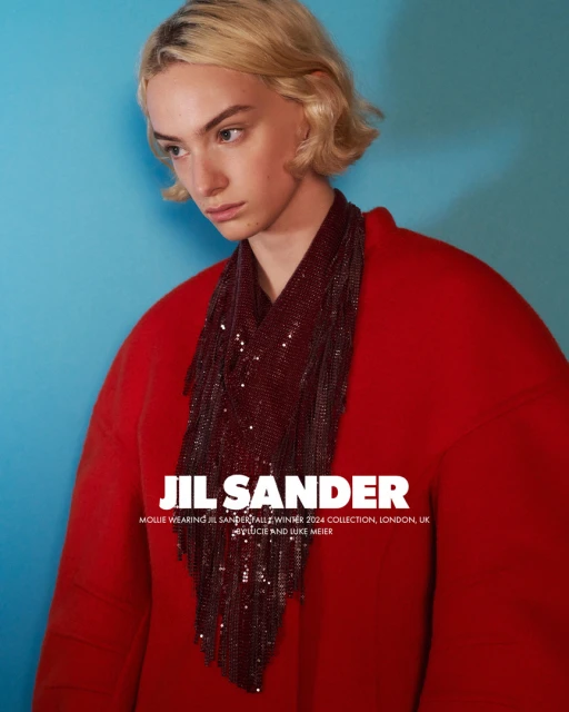 A model with short blonde hair poses in a studio wearing a red coat and beaded shawl against a turquoise background, fashion editorial style.