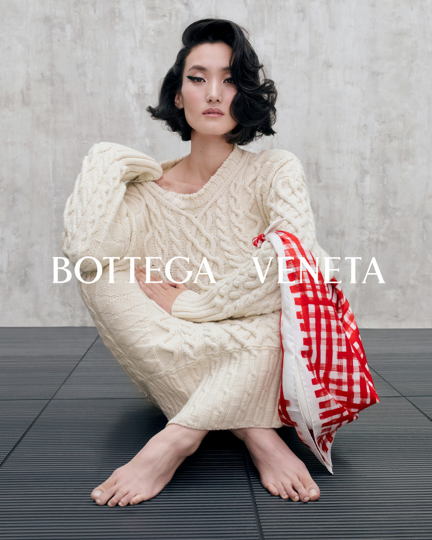 A woman in a cream-colored sweater dress and short dark hair, seated on a dark floor with a red and white checkered bag.