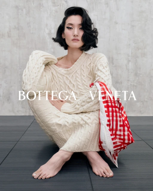 A woman in a cream-colored sweater dress and short dark hair, seated on a dark floor with a red and white checkered bag.