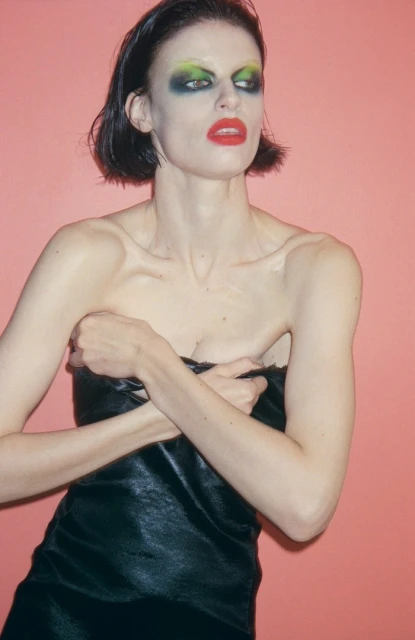 A woman with bold makeup poses against a pink background, wearing a black dress and crossing her arms for a fashion portrait.
