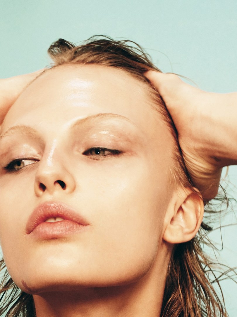 Close-up portrait of a fair-skinned woman with wet hair slicked back, pale pink lips, and hands on head, set against a light blue background.