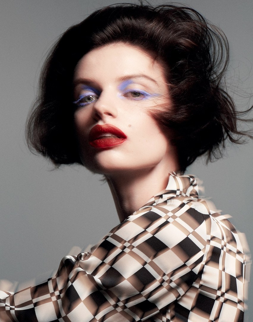 Close-up portrait of a fair-skinned woman with dark hair, bright red lipstick, and blue eyeshadow wearing a geometric patterned blouse.