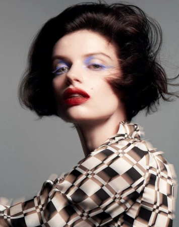 Close-up portrait of a fair-skinned woman with dark hair, bright red lipstick, and blue eyeshadow wearing a geometric patterned blouse.