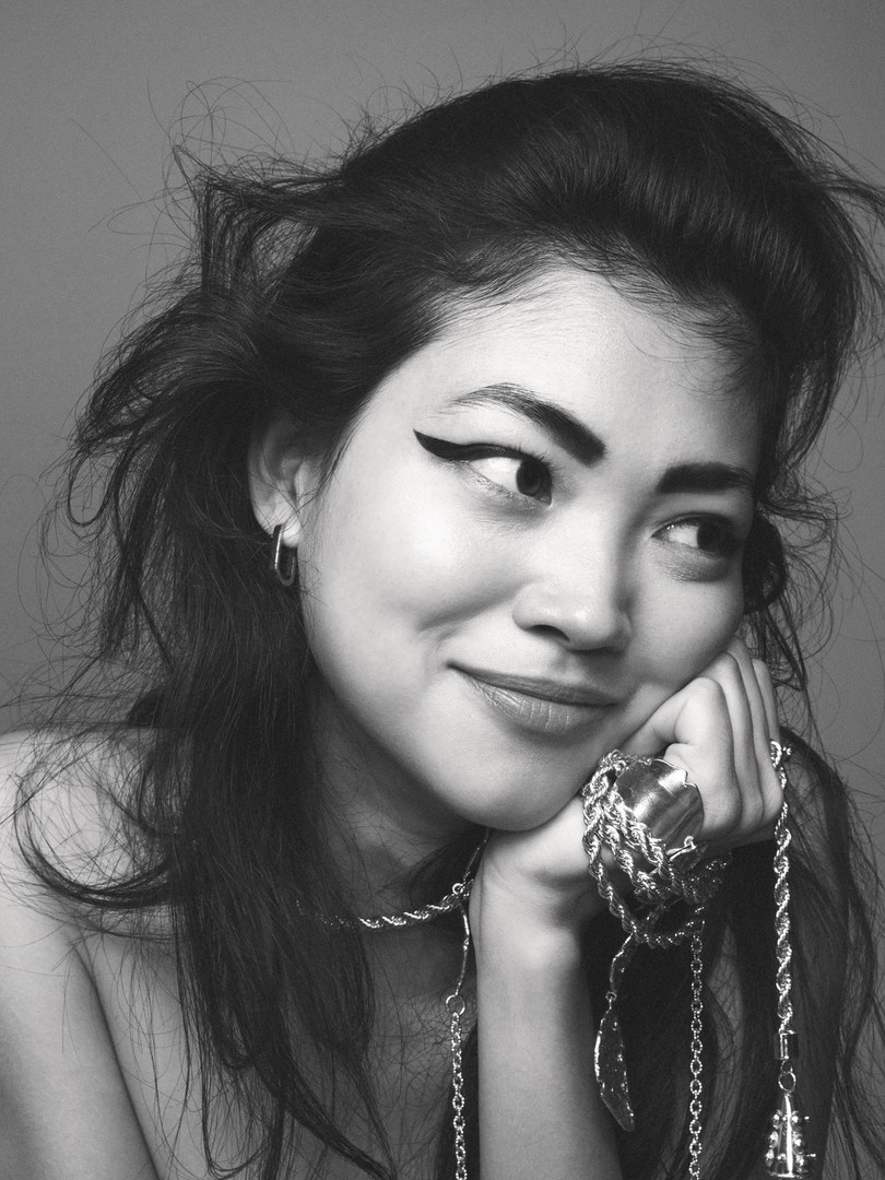 Close-up portrait of a woman with dramatic winged eyeliner resting her chin on her hand, wearing chunky silver jewelry.