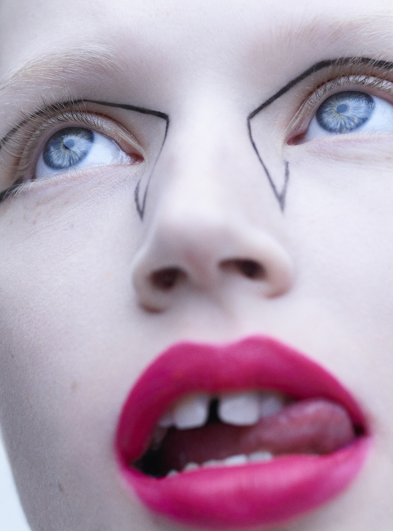 Close-up studio shot of a person with creative black liner, blue eyes, and bright pink lipstick; high-angle view focuses on facial features.