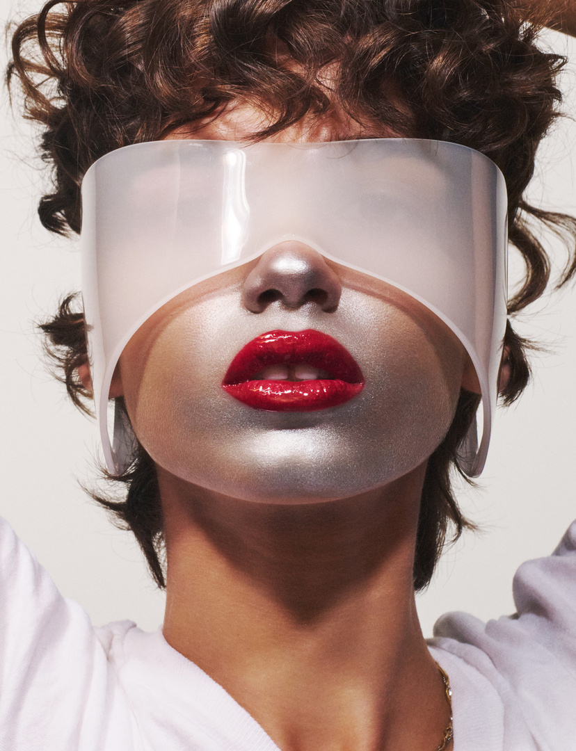 Close-up studio shot of person with silver face paint, red lips, and white visor against a plain background, fashion and beauty concept.