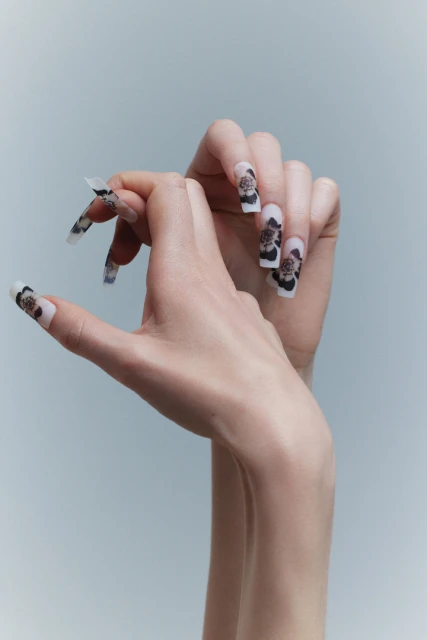 Close-up of two hands featuring long, square-shaped nails with a floral design against a soft blue background, showcasing beauty and style.