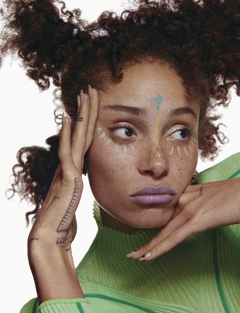 Close-up portrait of a young adult woman with textured brown hair, light-green top, and fantasy makeup on a white background.