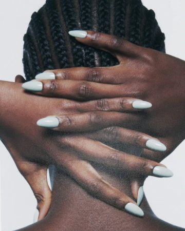 Close-up shot of a woman's hands with manicured nails covering the back of her neck with braided hair, set against a plain white backdrop.