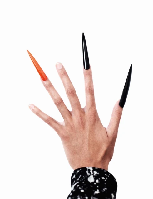 Hand with long, stiletto-shaped artificial nails, one orange, two black, against a stark white background, with patterned cuff visible.