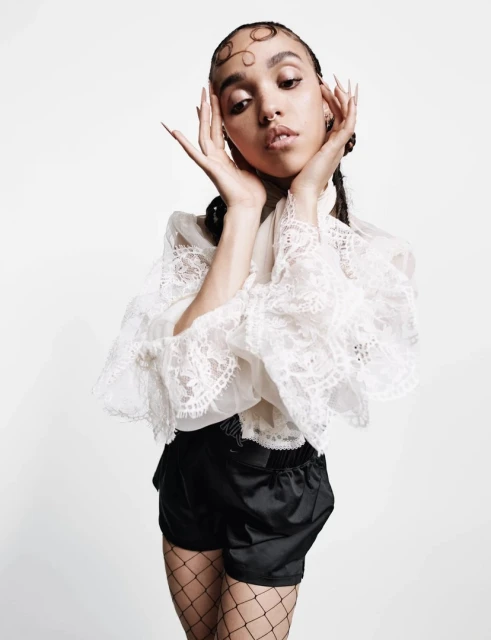 A woman with artistic hair and long nails poses in a white lace top and black shorts against a solid white backdrop in a studio setting.