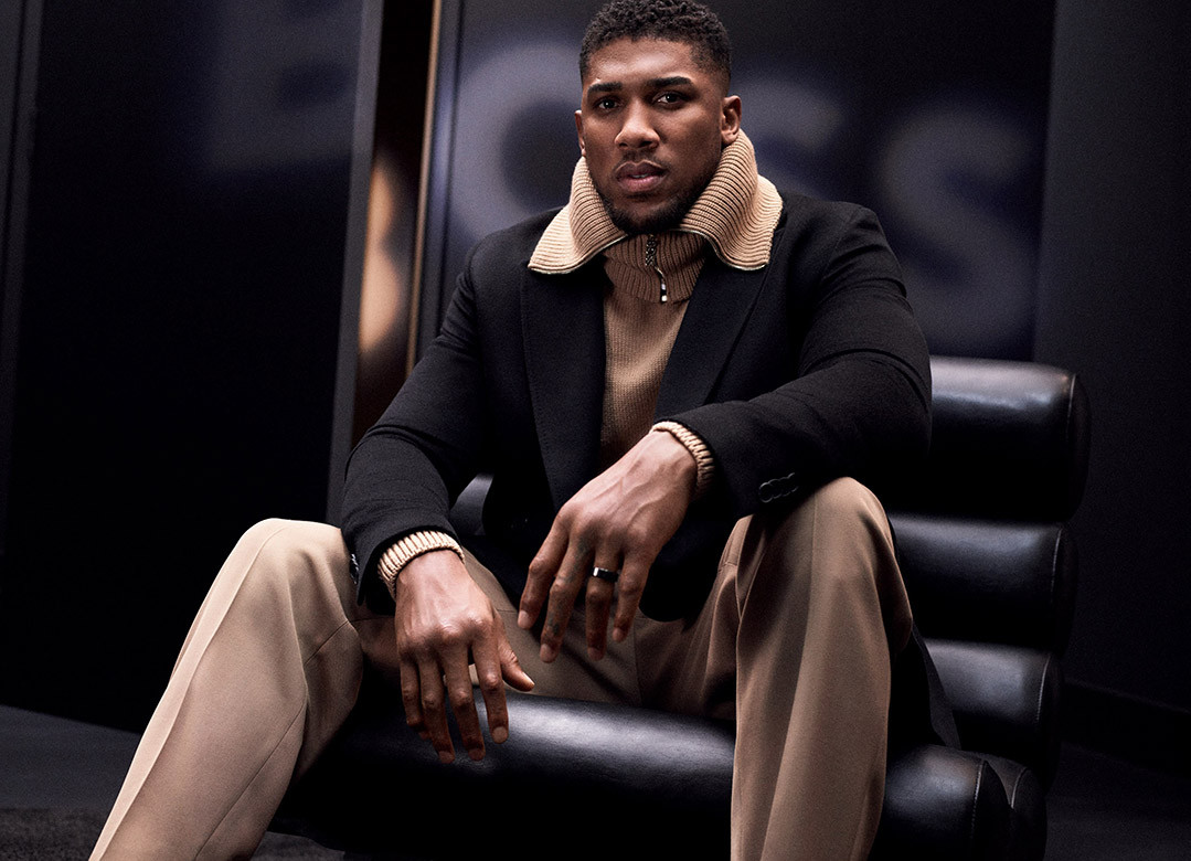Portrait of Anthony Joshua seated in a black chair, wearing a black jacket and beige pants, set against a dark background with 'BOSS' logo.