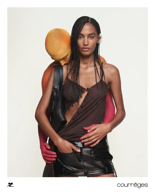 Fashion shot featuring a model in a sheer brown top and black skirt, with another person in orange body paint embracing her from behind.