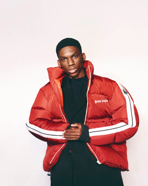A stylish African-American man poses in a red Palm Angels puffer jacket against a bright white backdrop, showcasing contemporary fashion.