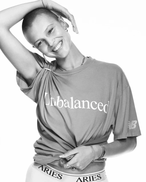 A smiling, bald woman poses in a studio, wearing a casual top and briefs with the brand name 'Aries'. The image is black and white.