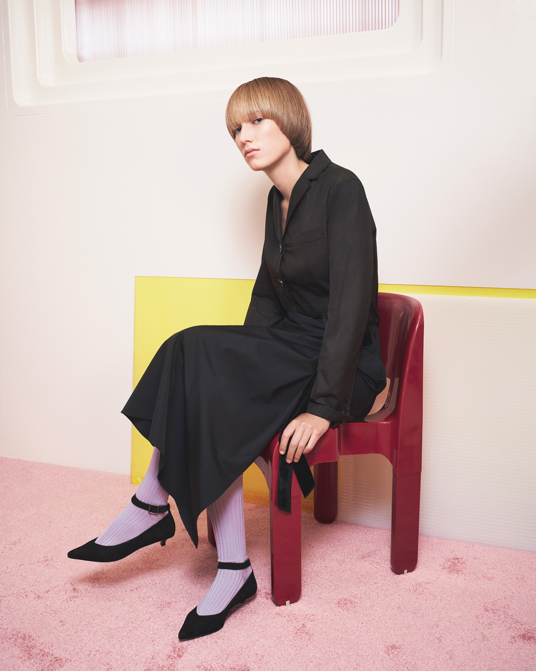 Young woman sits on a red plastic chair in a photo studio, wearing a black dress, black shoes, and light purple socks in a fashion editorial.