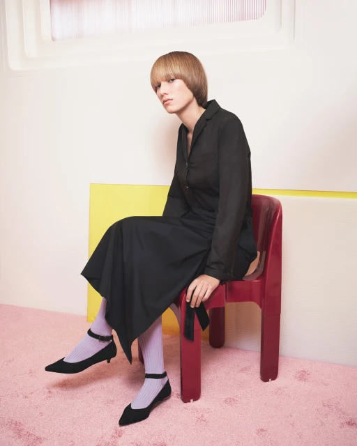 Young woman sits on a red plastic chair in a photo studio, wearing a black dress, black shoes, and light purple socks in a fashion editorial.