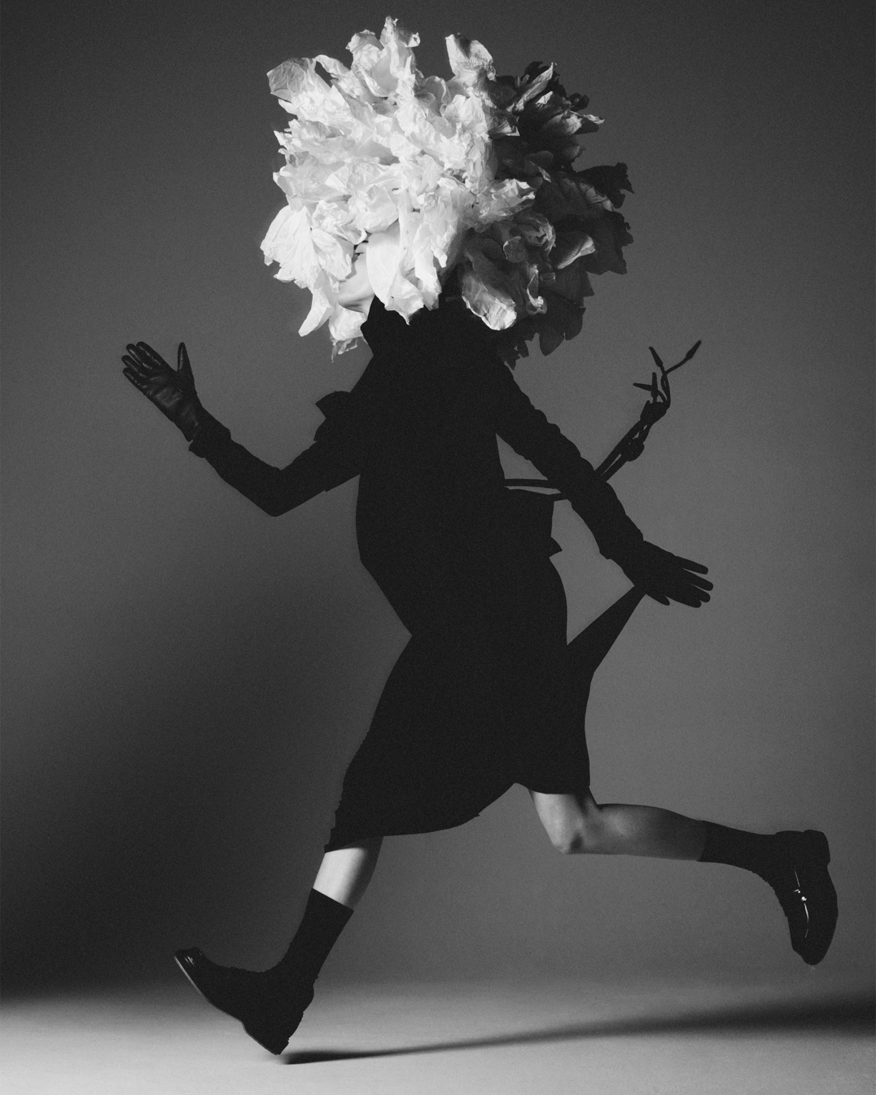 Monochrome fashion shot of a person in motion with a flower headpiece, running in a dark dress, gloves, and shiny shoes against a grey background.