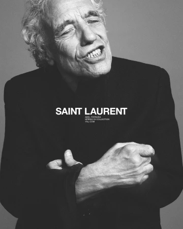Black and white portrait of an elderly man with curly hair wearing a dark jacket against a grey seamless background