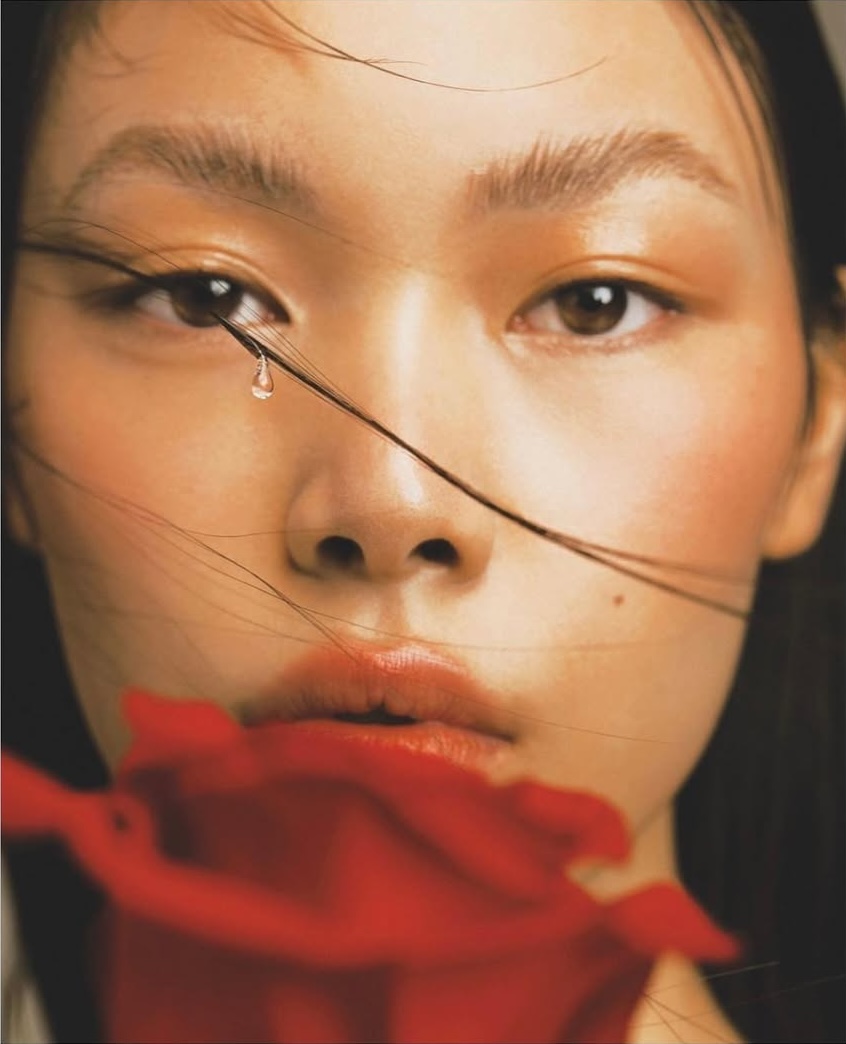 Close-up portrait of an Asian woman with a water droplet accent and a blurry red rose in the foreground, showcasing beauty and editorial.