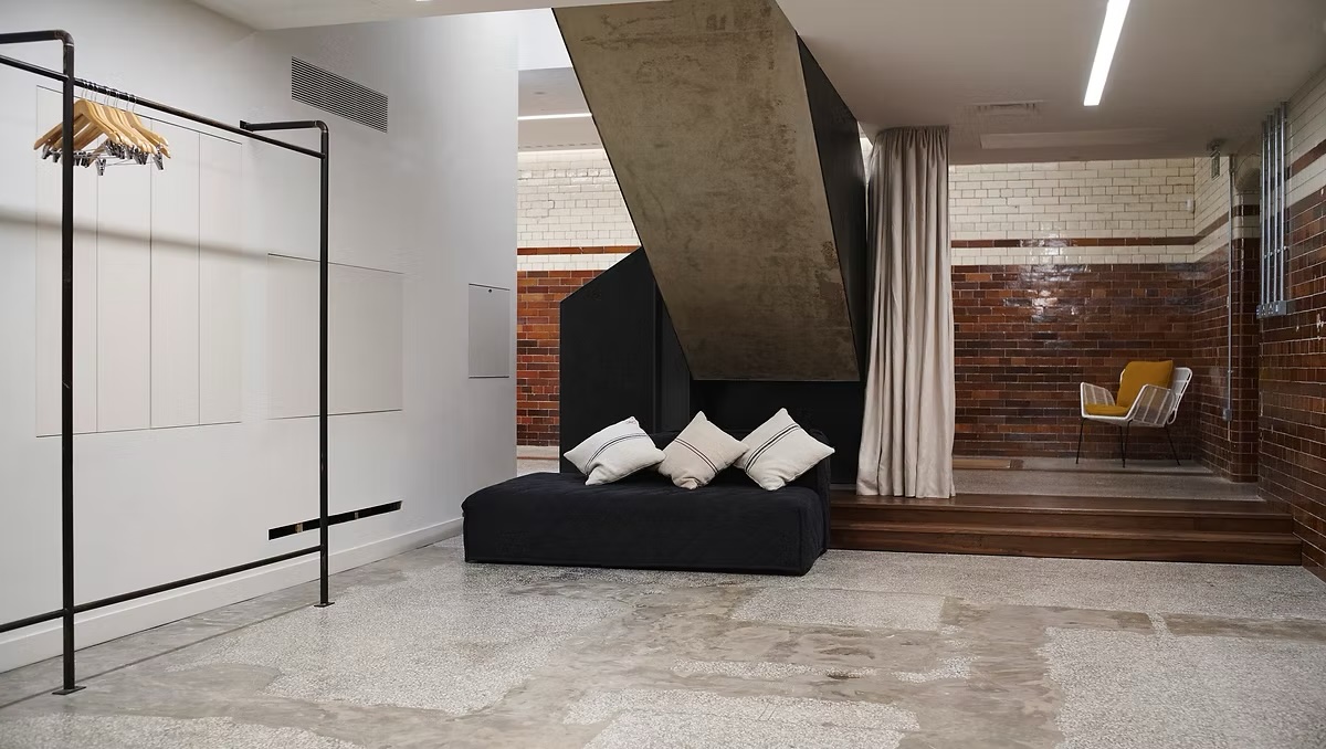 Modern interior showcasing a black daybed with white pillows, clothes rack, and staircase against a brown and white brick wall.