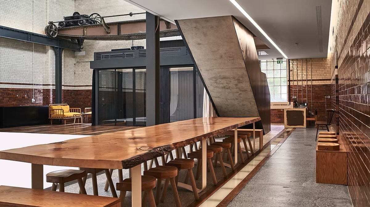 Spacious industrial interior with a long, live-edge wooden table, wooden stools, brick walls, and an overhead crane, creating a unique workspace.