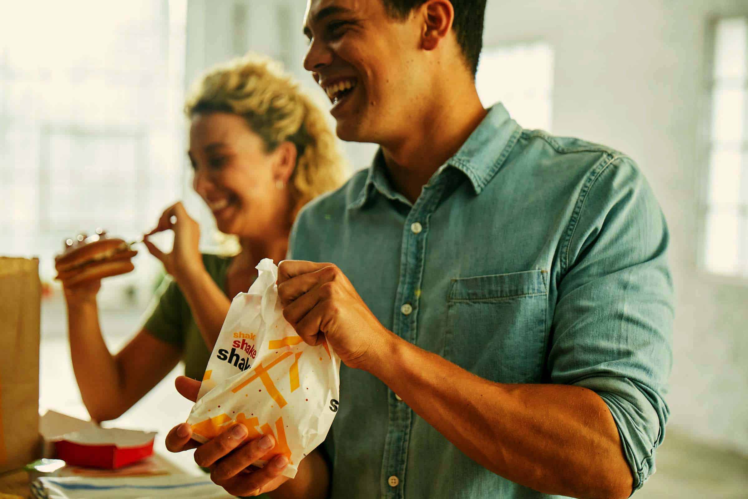 Two cheerful adults enjoy takeout food indoors near a bright window; one holds a bag of fries, the other a sandwich.