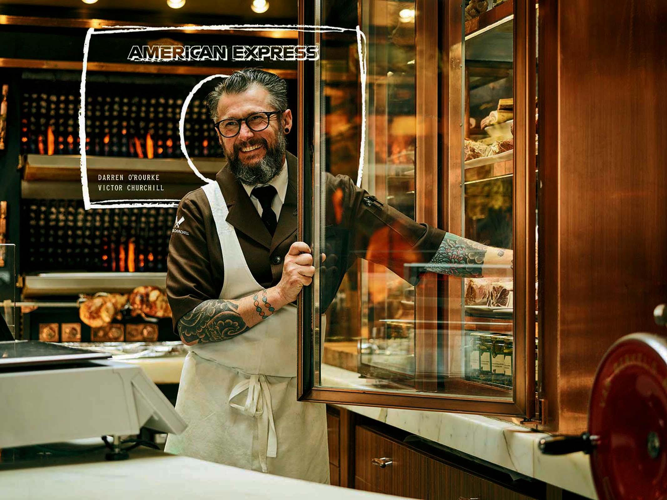 Chef with beard stands inside a restaurant near a glass display case and service counter with brand name and chef names visible.