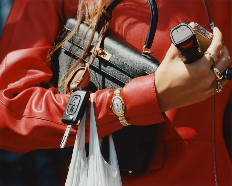 A woman in a red leather jacket holds a black purse, car keys, a gold lighter, and shopping bag, showcasing fashion and personal items.