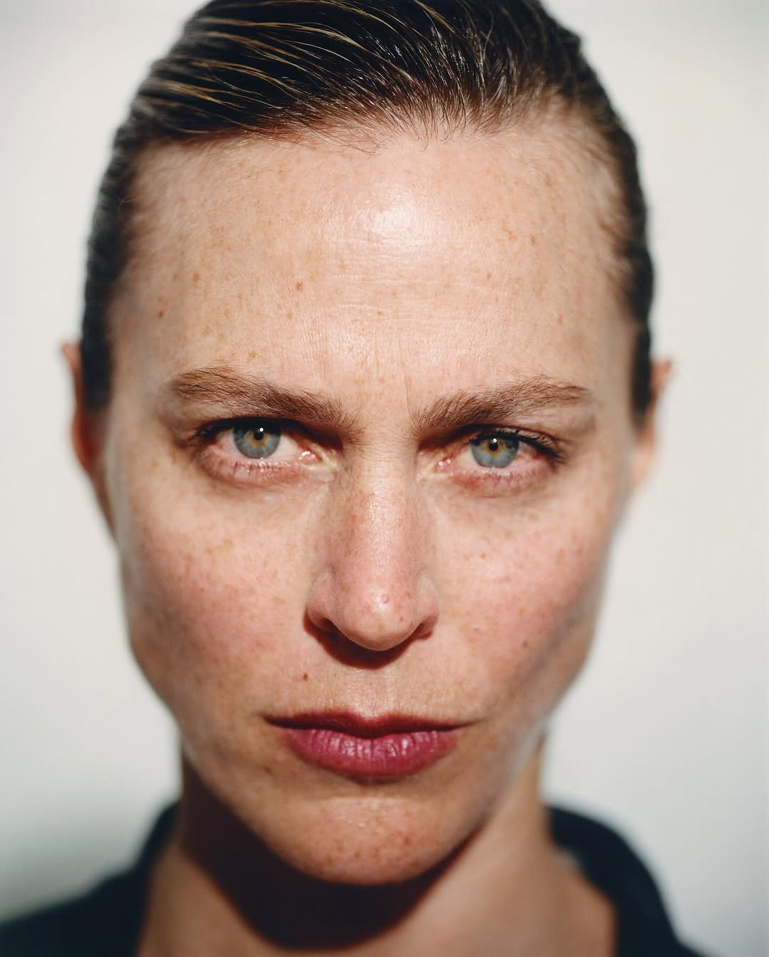 Close-up portrait of an adult woman with fair skin, green eyes, freckles, and slicked-back hair in a simple black top against a neutral backdrop.