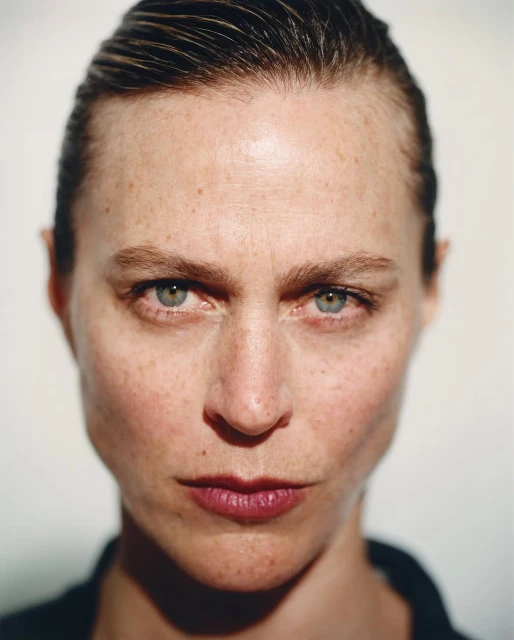 Close-up portrait of an adult woman with fair skin, green eyes, freckles, and slicked-back hair in a simple black top against a neutral backdrop.